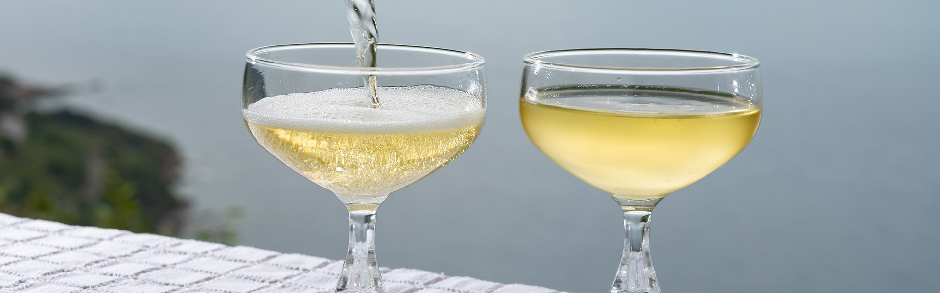 2487572597 Pouring of cold sparkling wine, cava or champagne in two coupe glasses on table with sea view