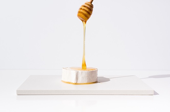2016261638 Pour honey over goat cheese, with wooden dropper on a white table. Appetizer. Mediterranean food