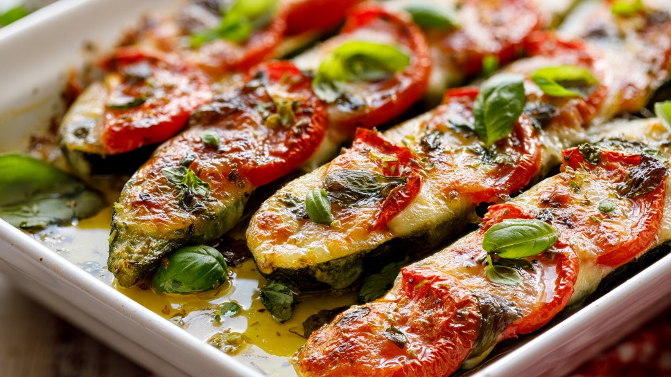 Roasted, stuffed zucchini with the addition of tomatoes, mozzarella cheese, fresh basil and olive oil (caprese salad) in a ceramic baking dish, close-up, top view. Nutritious and tasty vegetarian dish