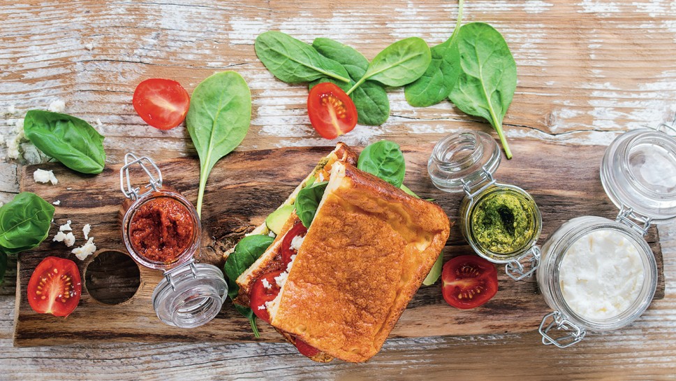 Low-carb gluten free Cloud bread veggie sandwich with spinach, avocado, feta cheese, tomatoes and pesto sauce, served on cutting board with jars of ingredients over wooden background. Top view