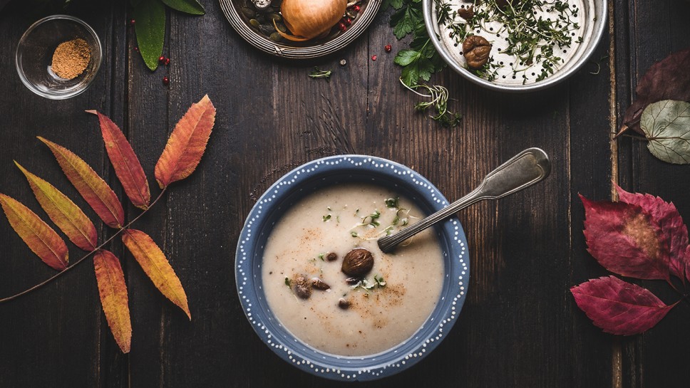 Vegetarian chestnuts creamy soup bowl on dark rustic kitchen table background with cooking pot ,ingredients and autumn leaves , top view. Healthy seasonal winter or autumn food and eating