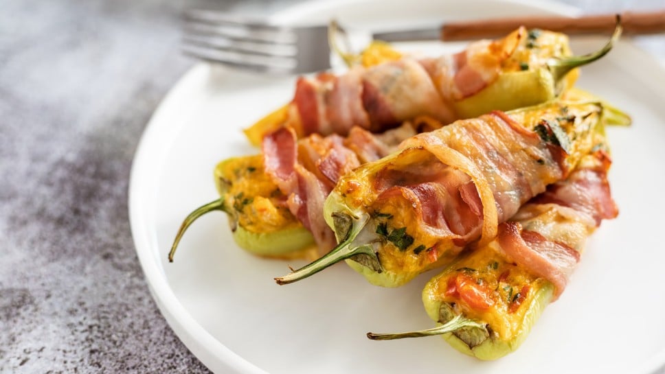 Green peppers stuffed with eggs, cheese, tomatoes, herbs wrapped in smoked bacon; baked vegetables and meat appetizer; warm salad served on a white round plate; finger food
