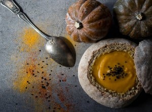 Squash soup with spices in fresh baked bread. Dark grey textured background. Acorn squash, silver spoon, bread bowl with soup.  