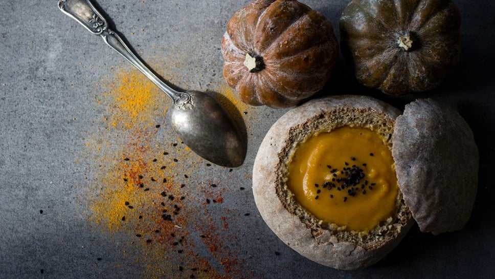 Squash soup with spices in fresh baked bread. Dark grey textured background. Acorn squash, silver spoon, bread bowl with soup.  