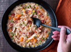 Woman’s hand with black metal spoon, Puerto Rican Chicken Rice Soup in a black stoneware bowl on a dark wood table, orange cloth napkin