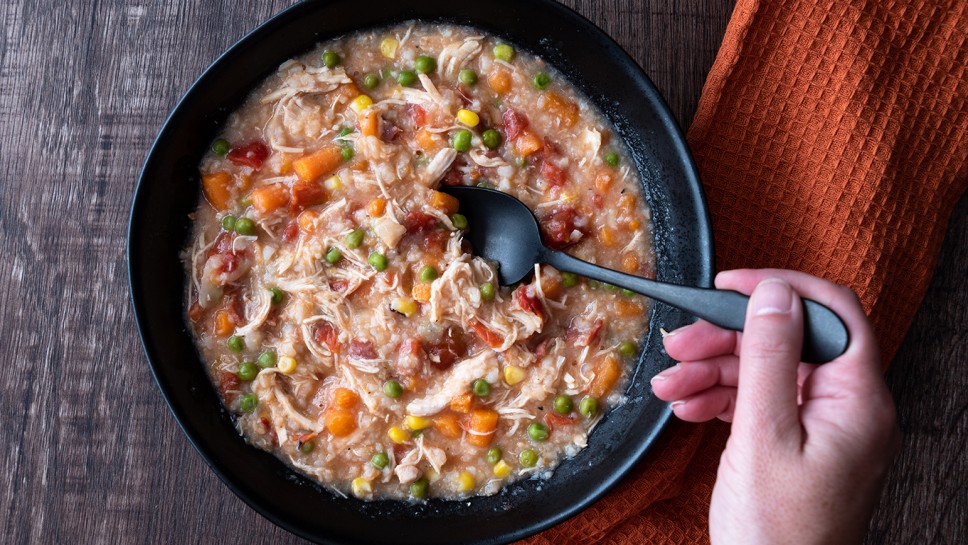 Woman’s hand with black metal spoon, Puerto Rican Chicken Rice Soup in a black stoneware bowl on a dark wood table, orange cloth napkin