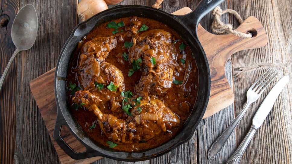 Chicken legs with brown onion sauce in a rustic cast iron pan on wooden table background. Top view with copy space