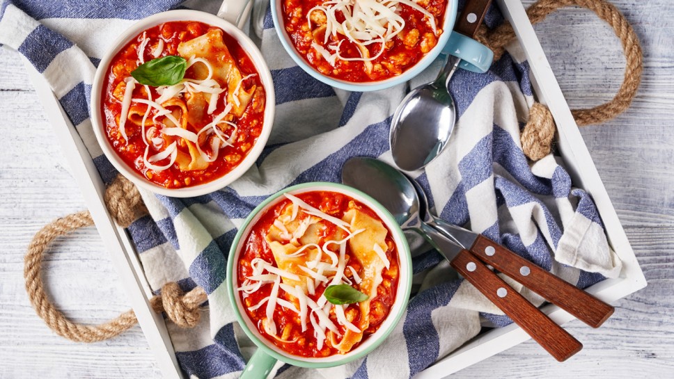 Healthy chicken lasagna soup with tomato, mozzarella, spices, basil served on a mugs with spoon on a white tray on a white wooden table, top view, close-up