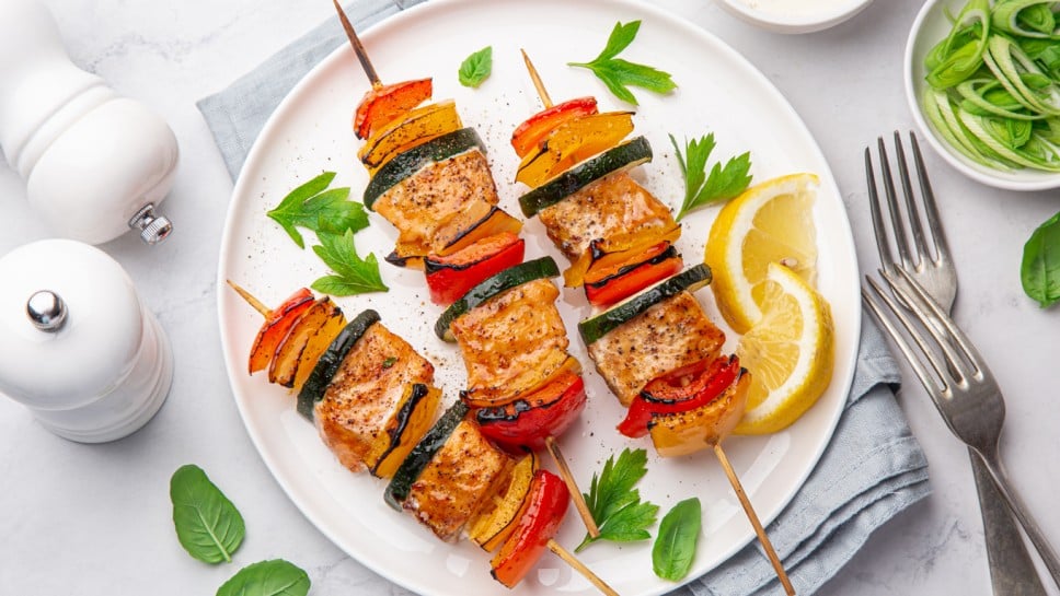 Grilled salmon and vegetables skewers on white plate, top view