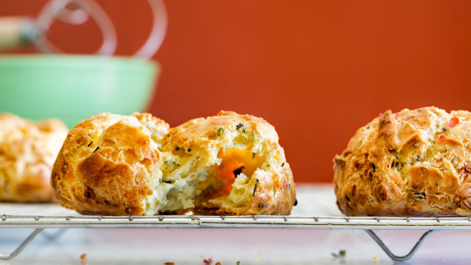 Cheese herb puff rolls on a cooling rack. One is pulled apart to show the inside.  French gougere. Green bowl with dough whisk. Close up view with copy space.