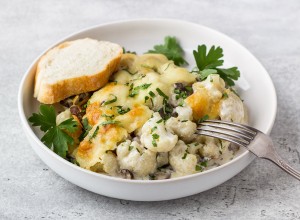 Baked cauliflower with mushrooms, cream and cheese sprinkled with parsley served with a slice of fresh bread and sour cream on a gray textured background, top view. Delicious homemade food