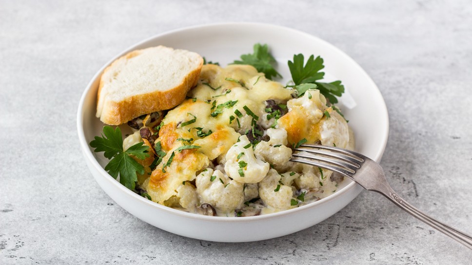 Baked cauliflower with mushrooms, cream and cheese sprinkled with parsley served with a slice of fresh bread and sour cream on a gray textured background, top view. Delicious homemade food