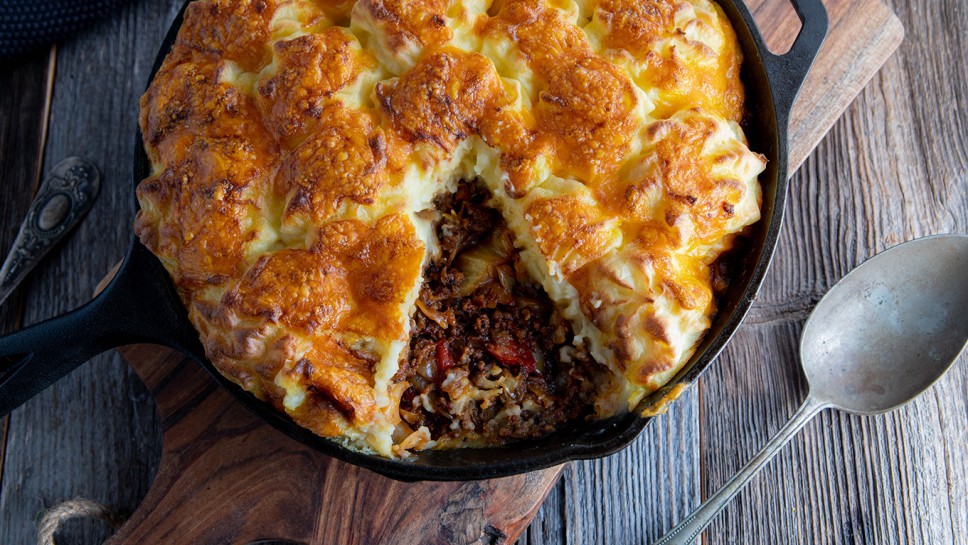 Savory pie with minced meat and cabbage. Topped with baked mash potato crust and cheddar cheese. Served in cast iron skillet on wooden table. Table top view