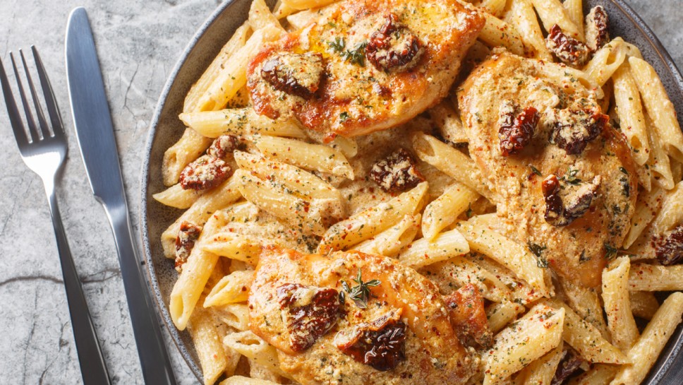 American Marry Me Chicken pasta with sun-dried tomatoes, cheese, herbs and aromatic creamy sauce close-up in a plate on a marble table. Horizontal top view from above