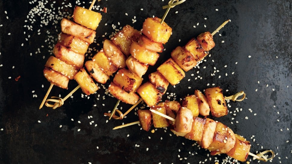 Grilled skewers with pineapple fruit and chicken meat  sprinkled with sesame seeds and spices on a black background, top view . Fruit and meat skewers