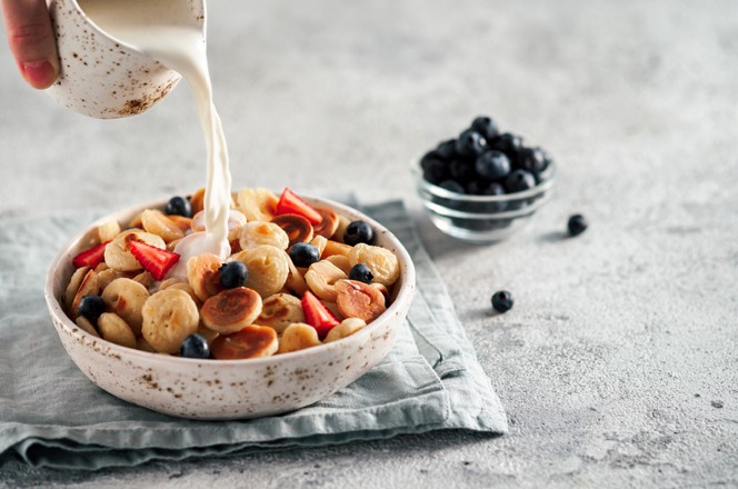 Trendy food - pancake cereal. Milk pouring on heap of mini cereal pancakes in bowl. Tiny cereal pancakes with berries in craft plate over gray cement background. Copy space for text or design.