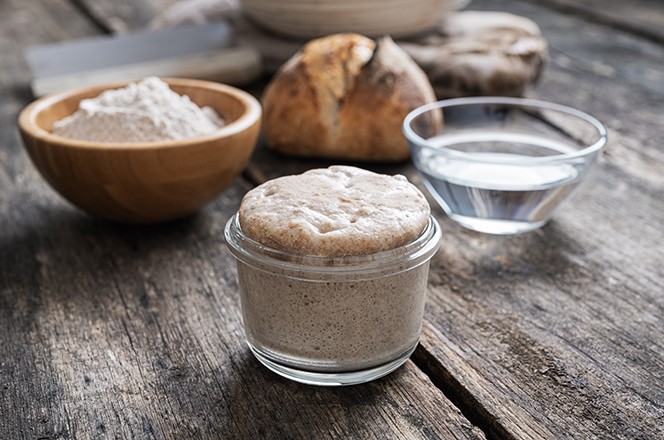 1752714791 Prepuna staklenka kvasa (sourdough starter) s domacom pogacom, bra nom i vodom u pozadini.