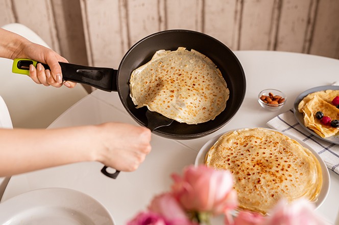 1761189836 Ruke mlade ene dr e tavu iznad kuhinjskog stola dok uzima vrucu, primamljivu palacinku kako bi je stavila na vrh druge hrpe palacinki na tanjuru.