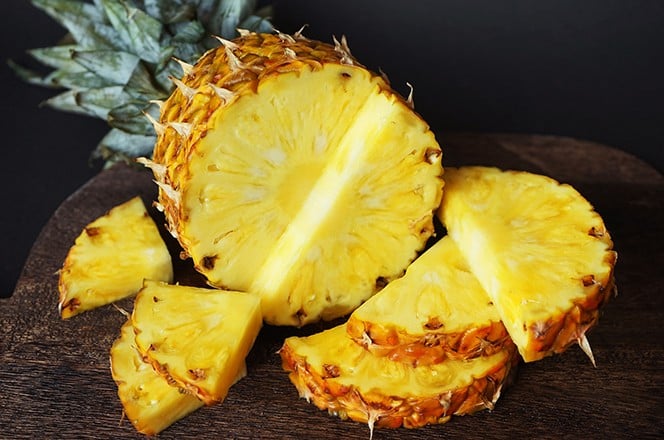 2470468107 Zreli socni ananas pored narezanih komada na drvenoj dasci, na tamnoj podlozi.