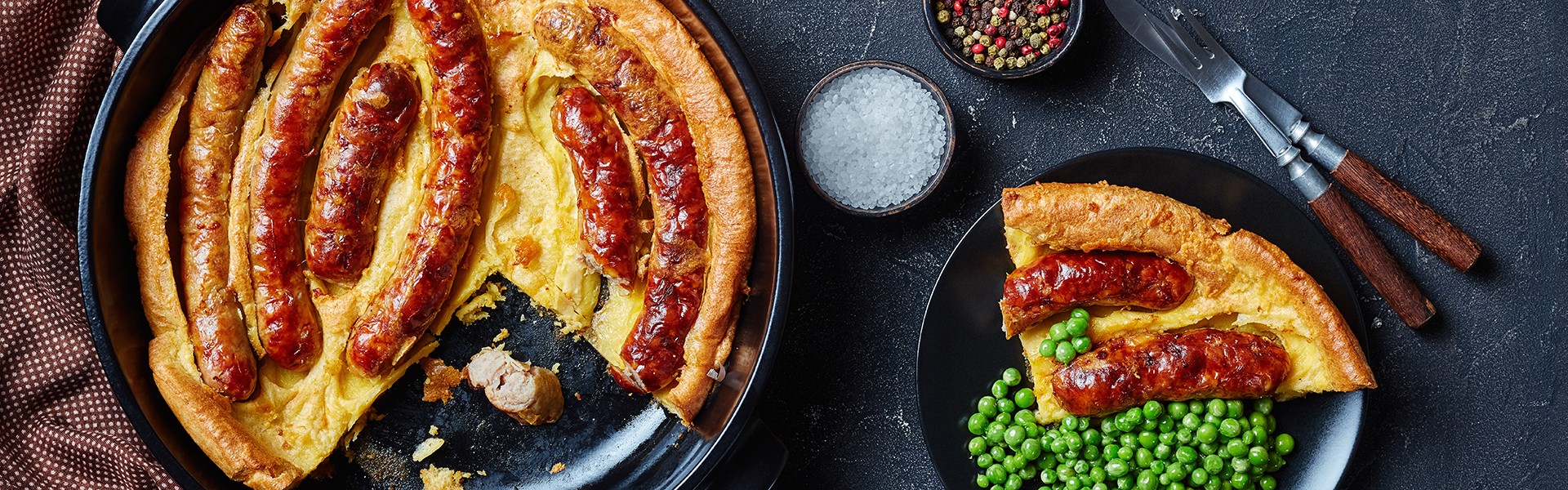 Toad in the hole, Sausage Toad, sausages in Yorkshire pudding batter in black ceramic baking dish and a portion on a plate with green peas and onion gravy,  view from above, flatlay