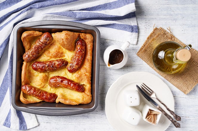 Toad in the hole of yorkshire pudding with roasted sausages served on a square cake pan with onion gravy, plate and cutlery on a white wooden background, horizontal view
